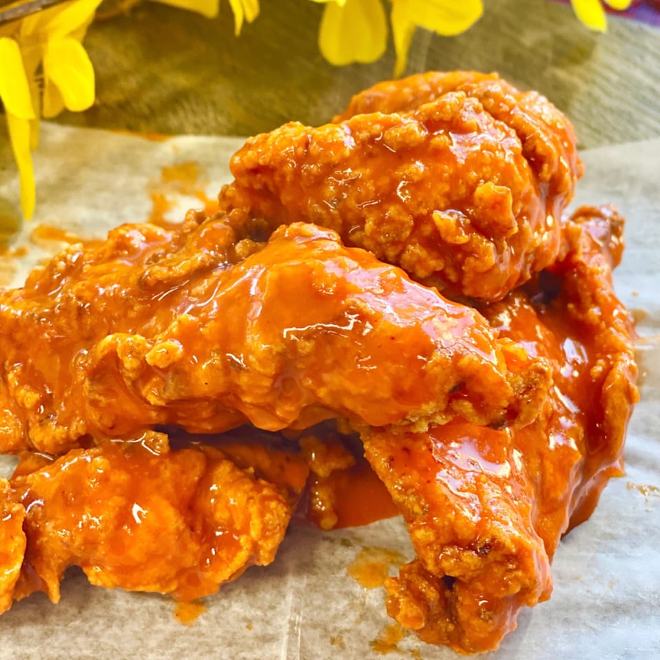 Close-up of Wingz's crispy, orange-glazed buffalo wings on parchment paper, with yellow flowers in the background.